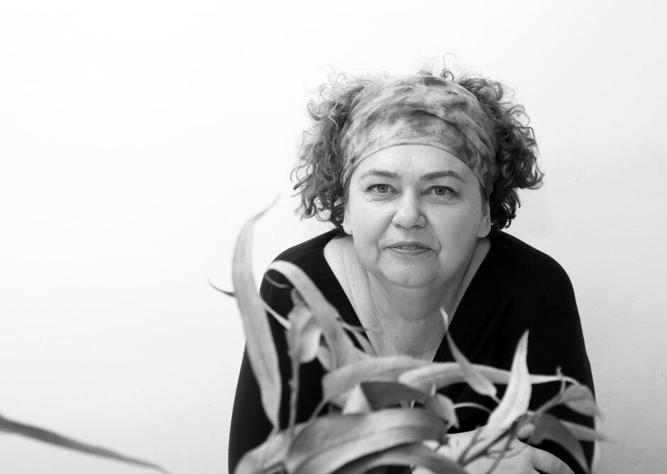 Na zdjęciu kobieta w opasce na głowie, przed nią roślina z długimi liśćmi.