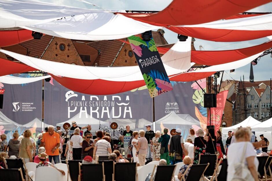 Na zdjęciu przedstawiona jest scena plenerowa przygotowana na wydarzenie „Parada – Piknik Seniorów” w Gdańsku. W centrum kadru widoczna jest duża scena, przed którą zebrała się grupa kilkudziesięciu seniorów. Niektórzy z nich tańczą, inni rozmawiają, a część odpoczywa na rozstawionych w pierwszym planie białych leżakach. Nad głowami uczestników rozwieszone są duże płachty materiału w kolorach czerwonym i białym, tworzące dekoracyjne zadaszenie, chroniące częściowo przed słońcem. Na tle sceny znajdują się dwa duże banery z nazwą wydarzenia: „Parada Piknik Seniorów”, logotypem Miasta Gdańsk oraz logotypem Regionalnego Centrum Wolontariatu w Gdańsku. Po bokach sceny zamontowano kolorowe flagi ozdobne z motywem liści tropikalnych w jaskrawych kolorach. W tle widać zabytkowe budynki starego miasta z czerwonymi dachami, charakterystycznymi dla Gdańska. Pogoda jest słoneczna, niebo częściowo zachmurzone. Całość obrazu oddaje atmosferę letniego, radosnego spotkania seniorów w przestrzeni miejskiej.
