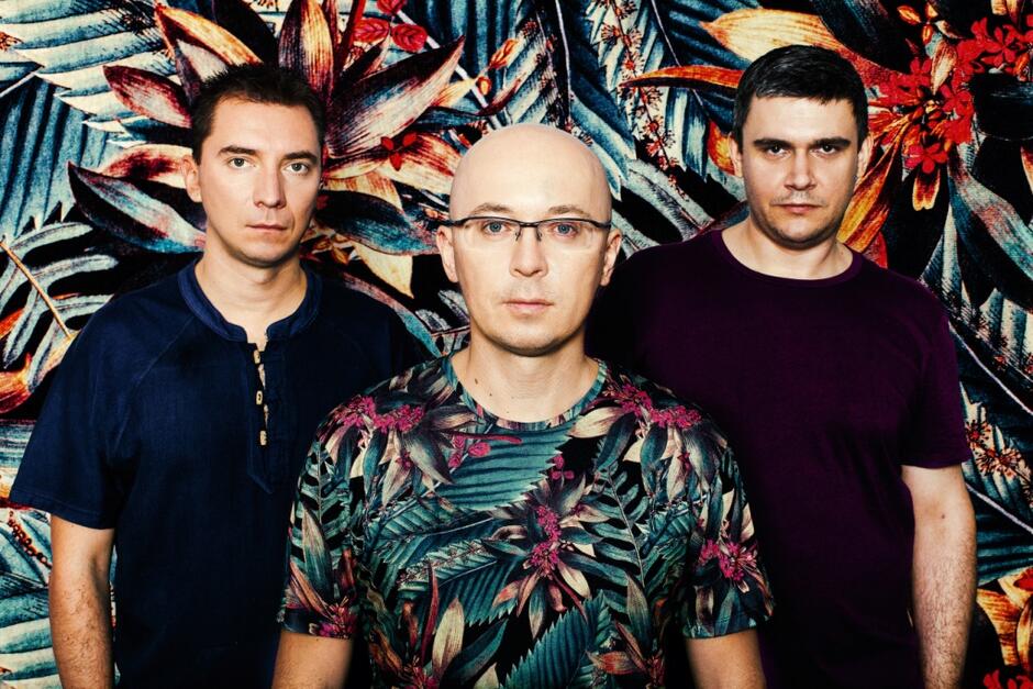Trzech mężczyzn stoi na tle intensywnie kolorowego, roślinnego wzoru – centralnie znajduje się łysy mężczyzna w okularach i koszulce w liście. Ich poważne wyrazy twarzy i skoordynowane pozy sugerują profesjonalny portret zespołu muzycznego