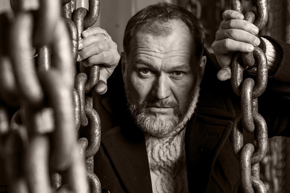 Mężczyzna z brodą i intensywnym spojrzeniem trzyma się masywnych, metalowych łańcuchów, jakby znajdował się w symbolicznym uwięzieniu. Zdjęcie w sepii dodaje dramatyzmu i podkreśla emocjonalny wyraz twarzy