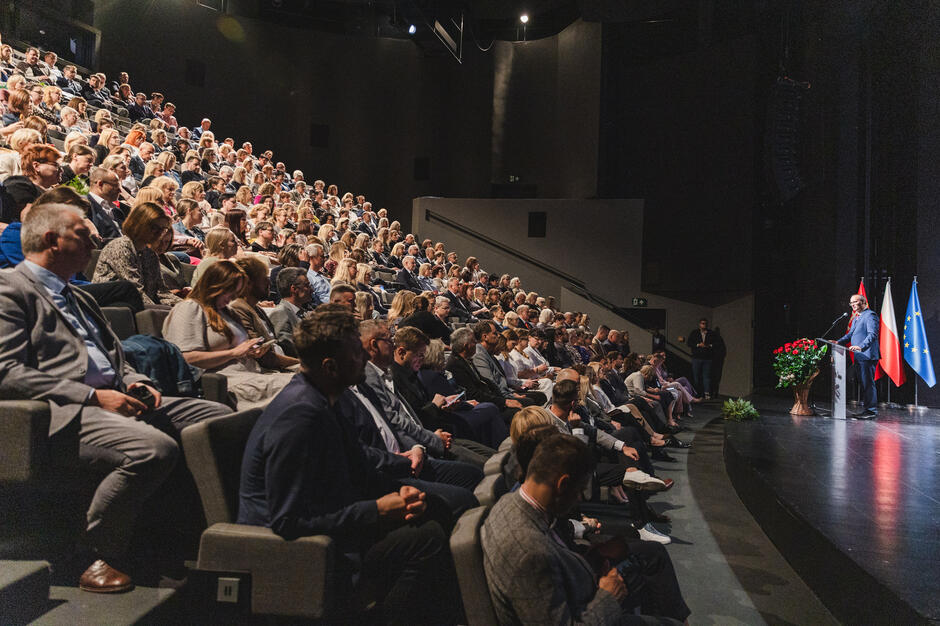 Mężczyzna przemawia na scenie z mównicy, na publiczności siedzi tłum słuchaczy.