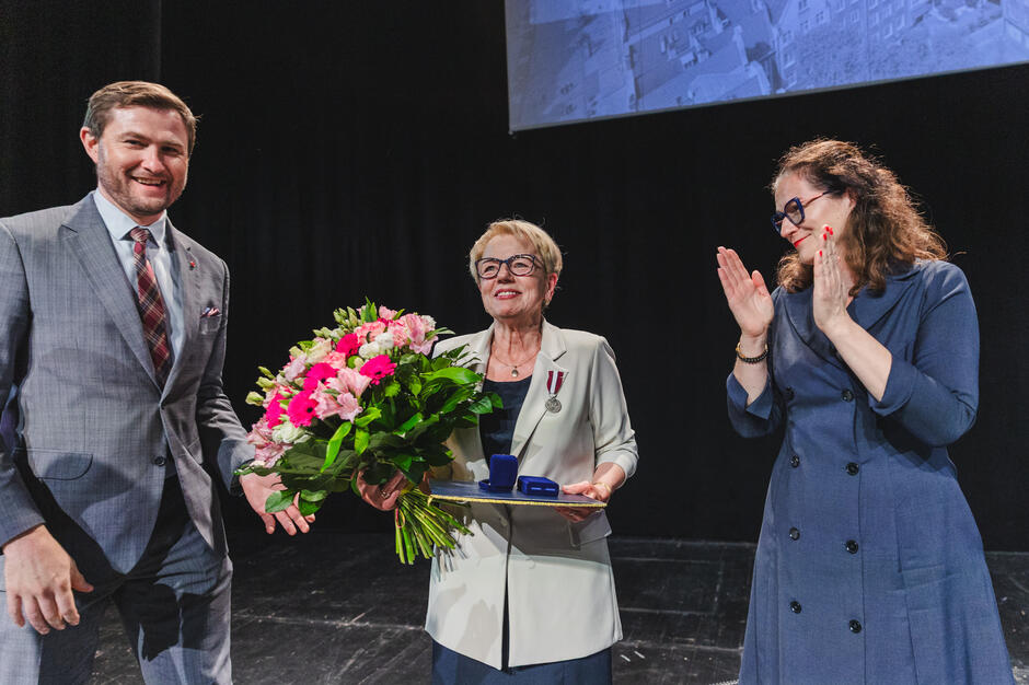 Trzy osoby na scenie podczas gali samorządowej. jedna kobieta ma przypięty medal, druga kobieta bije jej brawo, a mężczyzna wręcza bukiet kwiatów.