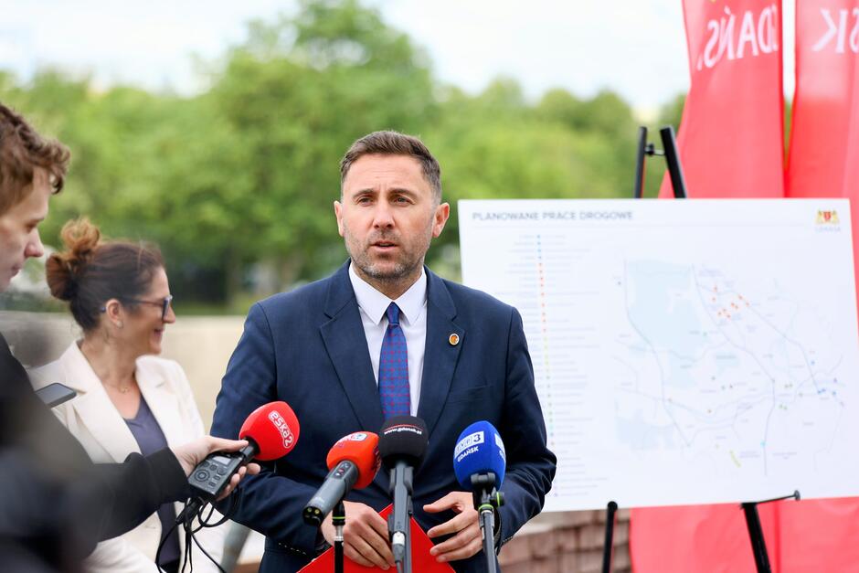 Na zdjęciu widoczna jest konferencja prasowa dotycząca planowanych prac drogowych w Gdańsku — mężczyzna w garniturze przemawia do mediów, a za nim stoi tablica z mapą inwestycji drogowych. W tle widać dziennikarzy z mikrofonami oraz banery z napisem „GDAŃSK”.