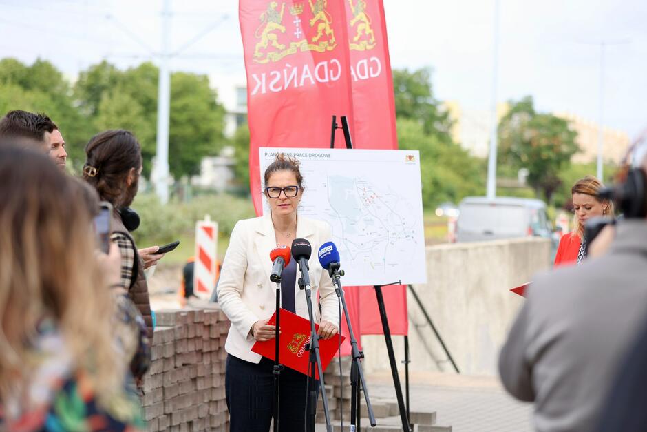 Na zdjęciu kobieta w jasnej marynarce przemawia podczas konferencji prasowej na temat planowanych prac drogowych w Gdańsku — za nią widać mapę inwestycji oraz czerwone banery z herbem miasta. Przed nią ustawione są mikrofony lokalnych mediów, a wydarzenie dokumentowane jest przez dziennikarzy i fotoreporterów.
