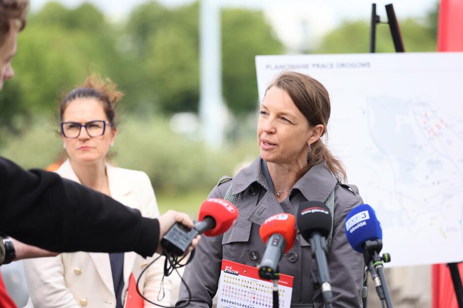 Na zdjęciu kobieta w szarym płaszczu wypowiada się przed mikrofonami mediów podczas konferencji dotyczącej planowanych prac drogowych w Gdańsku — za nią widoczna jest mapa inwestycji. W tle stoi inna kobieta w białej marynarce, uważnie przysłuchująca się wypowiedzi.