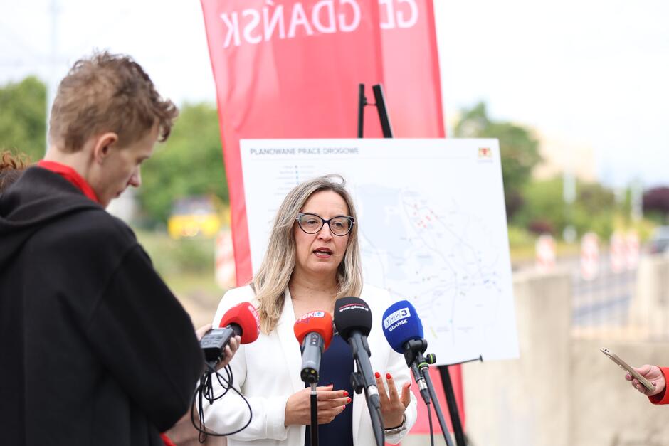 Na zdjęciu kobieta w okularach i białej marynarce przemawia do dziennikarzy podczas konferencji prasowej dotyczącej planowanych prac drogowych w Gdańsku. Za nią znajduje się mapa inwestycji oraz czerwony baner z herbem miasta, a przed nią ustawione są mikrofony kilku stacji telewizyjnych i radiowych.
