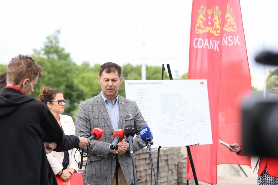 Na zdjęciu mężczyzna w szarej marynarce przemawia do mediów podczas konferencji prasowej poświęconej planowanym pracom drogowym w Gdańsku. W tle widoczna jest mapa inwestycji drogowych oraz czerwone banery z herbem i nazwą miasta, a obok stoją dziennikarze oraz inne osoby zaangażowane w wydarzenie.