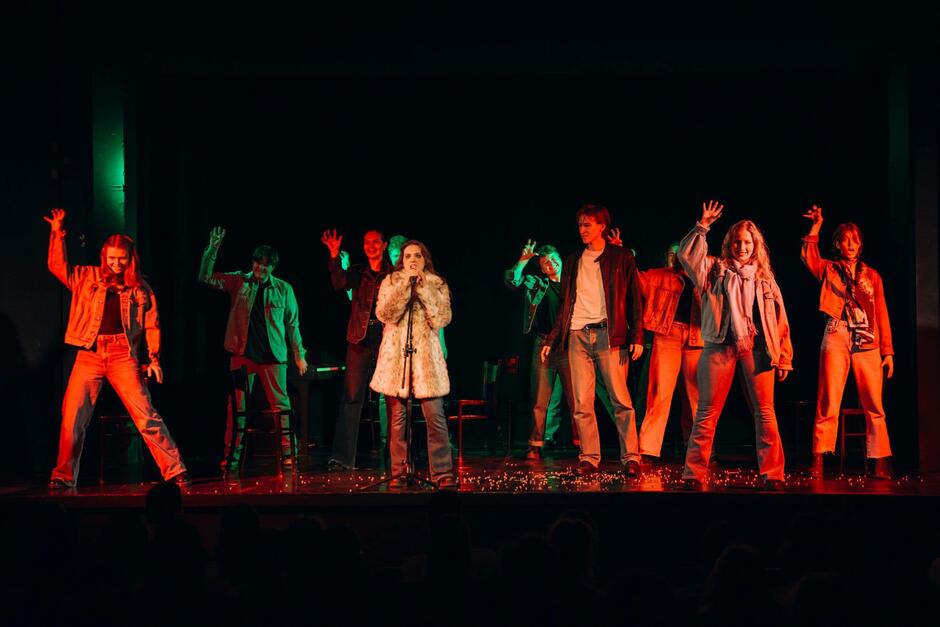 Zdjęcie przedstawia grupę młodych osób występujących na scenie teatralnej – w centrum stoi kobieta w futrzanym płaszczu, śpiewająca do mikrofonu, a pozostali wykonują zsynchronizowany gest z uniesionymi rękami. Scena jest oświetlona czerwono-zielonym światłem, a publiczność w cieniu obserwuje występ.