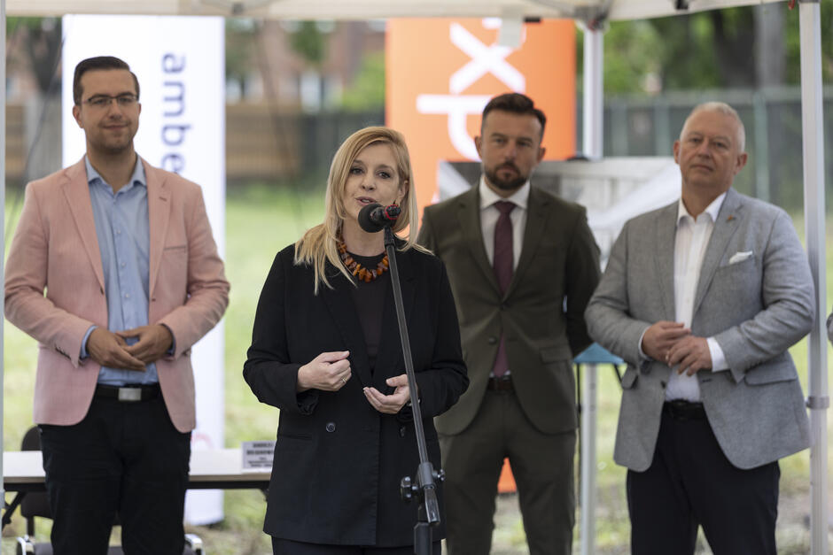 Kobieta przemawia do mikrofonu w trakcie uroczystości, a za nią stoją partnerzy projektu. Atmosfera spotkania sugeruje ważne ogłoszenie dotyczące przyszłości inwestycji