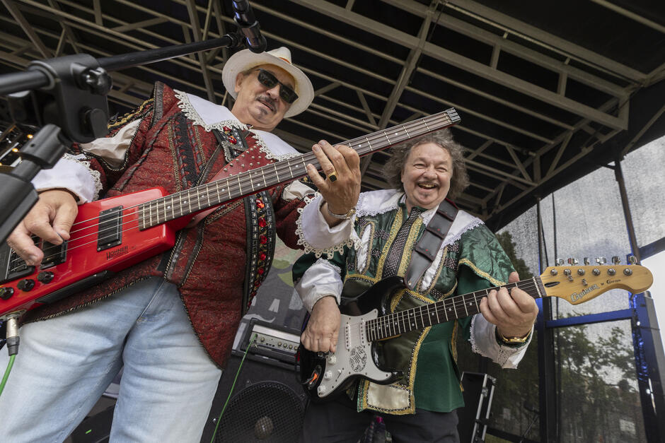 Na zdjęciu dwóch uśmiechniętych muzyków gra na gitarach na scenie plenerowej – jeden na czerwonym basie, drugi na czarnej gitarze elektrycznej. Ubrani są w kolorowe, stylizowane stroje przypominające renesansowe kostiumy, co nadaje występowi radosnego i teatralnego charakteru.
