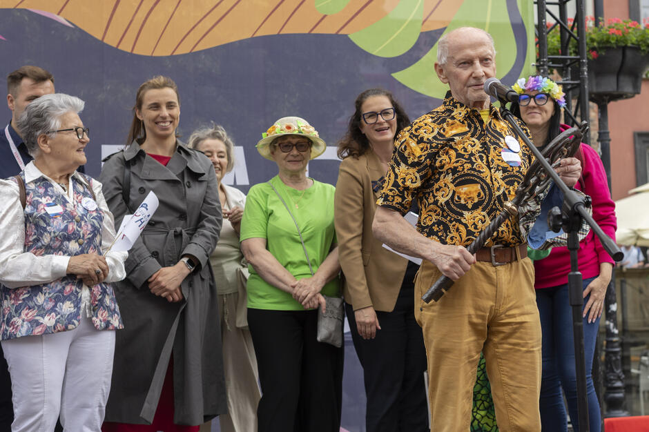 Na zdjęciu widać grupę uśmiechniętych seniorów i seniorek stojących na scenie podczas wydarzenia plenerowego – jeden z mężczyzn przemawia do mikrofonu, trzymając ozdobne berło. Tło zdobi kolorowa grafika, a atmosfera jest radosna i pełna pozytywnej energii.

