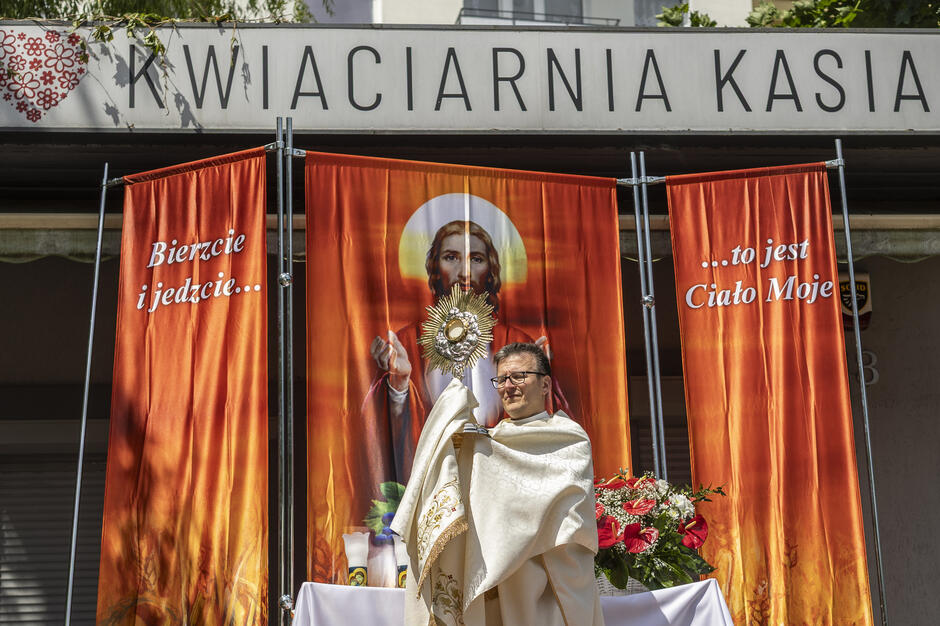 Ksiądz z monstrancją przy ulicznym ołtarzu, nad nim szyld z napisem Kwiaciarnia Kasia