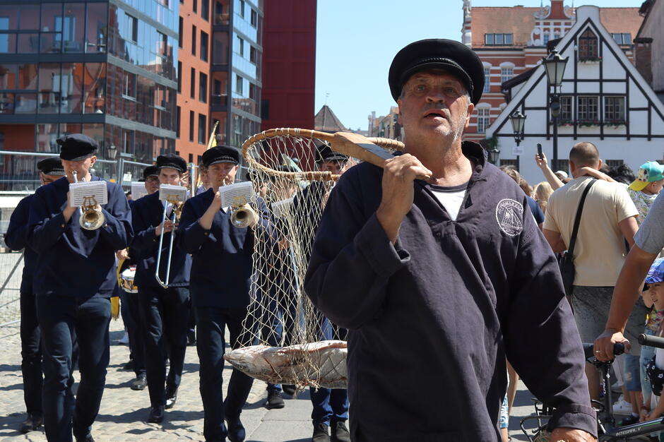 Idą kaszubscy rybacy z czarnych czapkach z daszkiem. Pierwszy z nich niesie na ramieniu podbierak, a w nim dużą rybę
