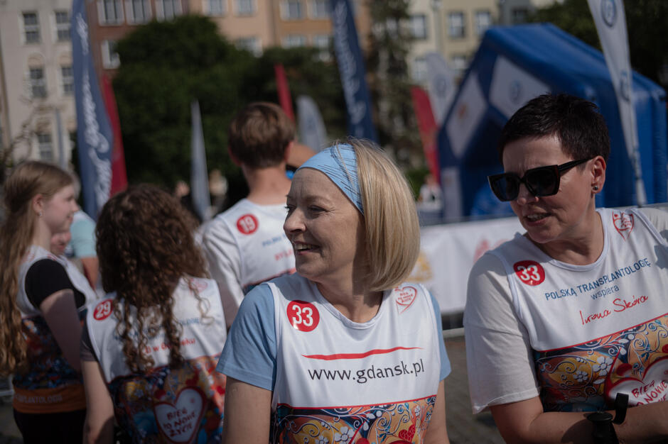 Uczestniczki wydarzenia „Bieg po Nowe Życie”, Iwona i Ilona, ubrane w oficjalne koszulki z numerem startowym 33. Na koszulce widoczne logo Gdańska i napis „www.gdansk.pl”. Obie kobiety uśmiechnięte, gotowe do startu, otoczone innymi uczestnikami – w tle flagi partnerów i charakterystyczne zabudowania Głównego Miasta.