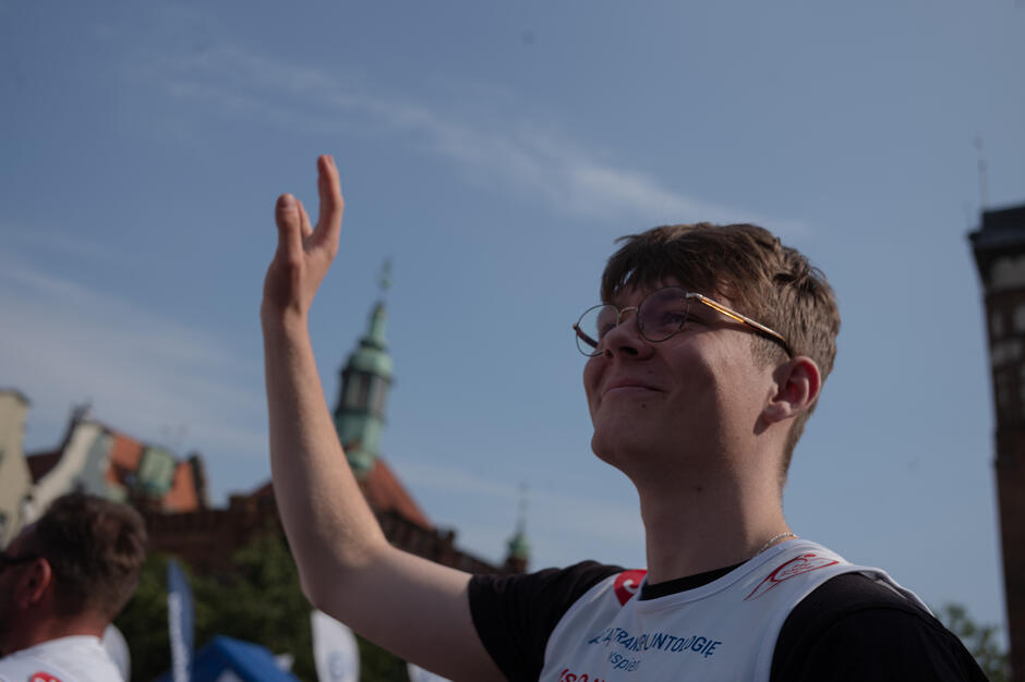 Tymoteusz, reprezentujący młodych uczestników wydarzenia „Bieg po Nowe Życie” bierze udział w rozgrzewce – unosi ramię w górę z uśmiechem na twarzy. Ubrany w oficjalną koszulkę z numerem startowym i okularami, na tle nieba i wieżyczek historycznych gdańskich kamienic. W tle widoczne są także flagi partnerów i inni uczestnicy wydarzenia.
