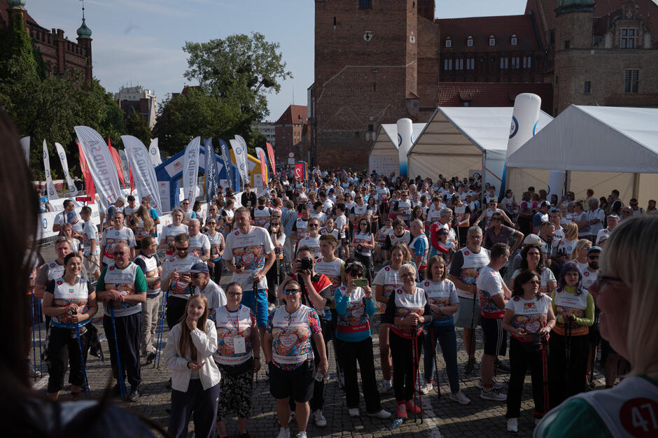 Tłum uczestników wydarzenia „Bieg po Nowe Życie” zgromadzony na starcie przy Zbrojowni w Gdańsku. Widoczna różnorodność wiekowa uczestników – od młodzieży po seniorów – wszyscy ubrani w charakterystyczne, kolorowe koszulki biegu z sercem. W tle banery sponsorów i namioty organizatora. Atmosfera skupienia i radości przed rozpoczęciem marszu nordic walking.
