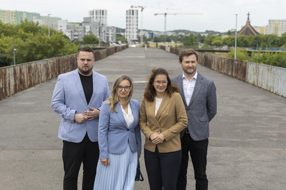 Cztery osoby - dwie kobiety i dwóch mężczyzn pozuje do zdjęcia na kładce dla pieszych w mieście.