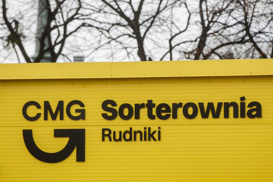 Na zdjęciu widoczna jest żółta elewacja budynku z czarnym napisem „CMG Sorterownia Rudniki” oraz charakterystycznym logotypem. W tle widać bezlistne drzewa, co sugeruje, że zdjęcie wykonano jesienią lub zimą.