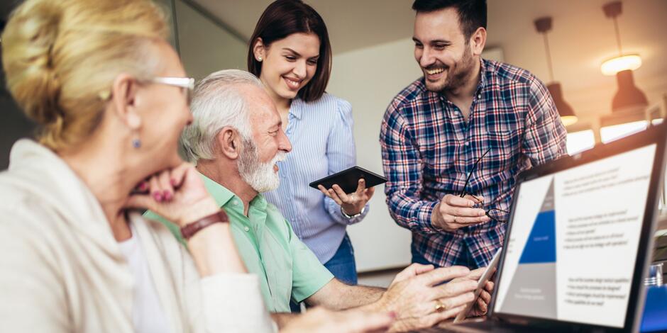 Na zdjęciu widać międzypokoleniową grupę czterech osób podczas wspólnej pracy lub warsztatów. Dwie starsze osoby – kobieta i mężczyzna – siedzą przy stole, skupione na ekranie laptopa i tablecie. Starszy mężczyzna z siwą brodą nosi zieloną koszulę i uśmiecha się, trzymając tablet. Starsza kobieta z blond włosami spiętymi w kok opiera się na dłoni i patrzy z zainteresowaniem. Obok nich stoją dwie młodsze osoby – kobieta i mężczyzna – oboje uśmiechnięci, zaangażowani w rozmowę. Młoda kobieta w niebieskiej bluzce trzyma tablet, a młody mężczyzna w koszuli w kratę trzyma okulary i mówi z entuzjazmem. Na pierwszym planie, po prawej stronie, częściowo widoczny jest ekran laptopa z otwartą prezentacją, zawierającą tekst i wykres w kolorze niebieskim.

Cała scena odbywa się w jasnym, nowoczesnym wnętrzu z widocznymi lampami sufitowymi w tle. Atmosfera jest przyjazna, zaangażowana i pełna współpracy.