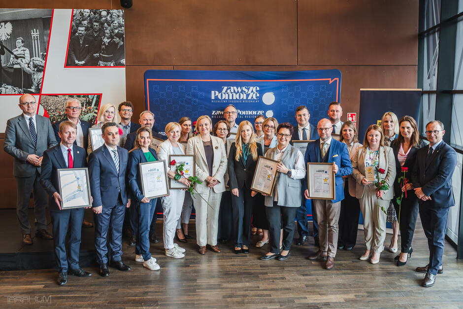 Zdjęcie grupowe wykonane w pomieszczeniu podczas oficjalnej uroczystości. Na fotografii znajduje się około dwudziestu elegancko ubranych osób – kobiet i mężczyzn – ustawionych w kilku rzędach. Wszyscy patrzą w kierunku aparatu, większość uśmiecha się. Osoby na pierwszym planie trzymają w rękach nagrody – prostokątne, oprawione dyplomy lub statuetki – oraz pojedyncze czerwone róże. Ubrani są w stroje biznesowe, takie jak garnitury, marynarki i eleganckie kostiumy.