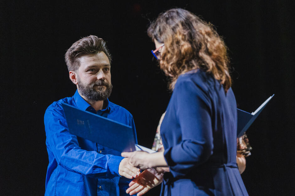 Mężczyzna w niebieskiej koszuli odbiera dokument z rąk kobiety w granatowym stroju podczas oficjalnej uroczystości. Sytuacja ma charakter formalny, prawdopodobnie związany z wręczeniem stypendium lub nagrody