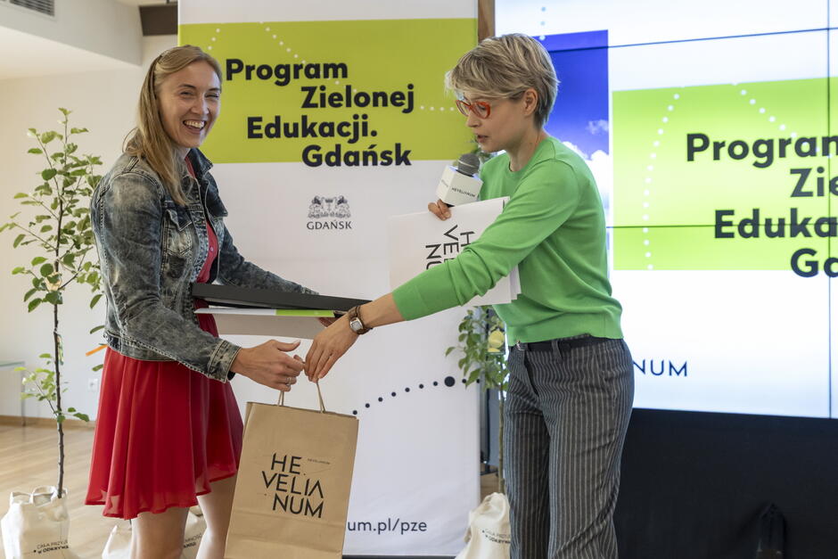 Na zdjęciu Karolina Kętrzyńska z Przedszkola nr 61 odbiera nagrodę, odznakę, certyfikat i dyplomy podczas finału Programu Zielonej Edukacji w Gdańsku. Wręczenia dokonuje przedstawicielka organizatora z Hevelianum, trzymająca mikrofon i materiały promocyjne.