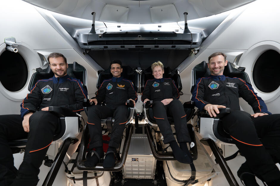 Czwórka astronautów na pokładzie rakiety - trzech mężczyzn i jedna kobieta, siedzą na fotelach i uśmiechają się.