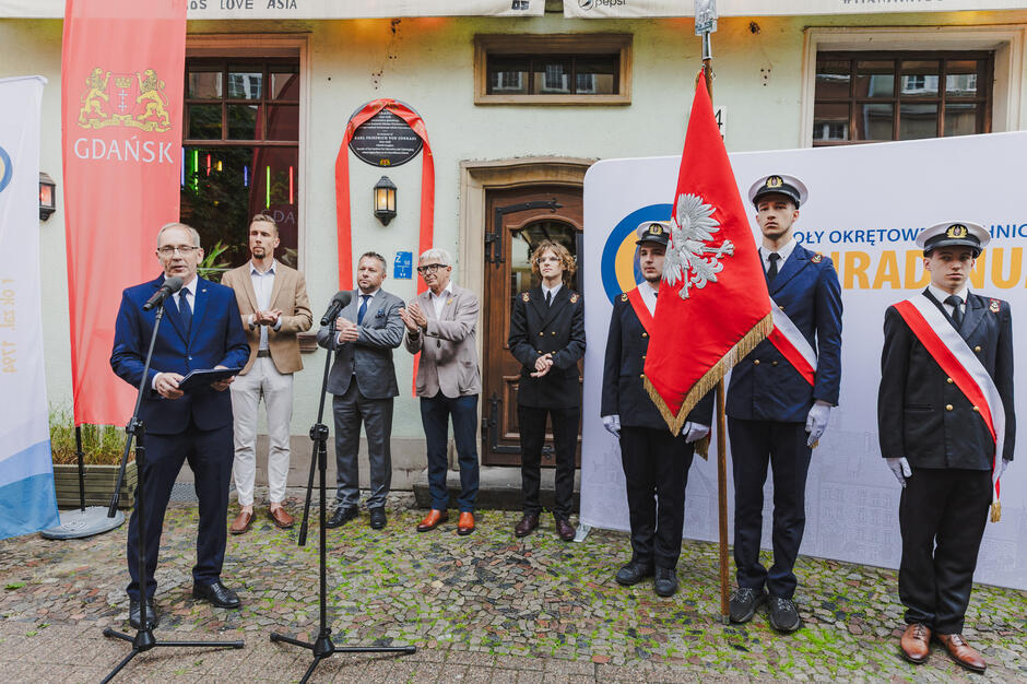 Poczet sztandarowy szkoły oraz kilku mężczyzn stoją pod kamienica, na której wisi tablica pamiątkową z czerwoną szarfą. jeden mężczyzna przemawia do mikrofonu.
