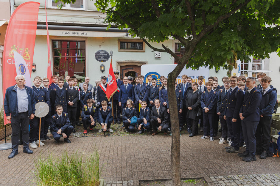 Uczniowie szkoły mundurowej pozują do zdjęcia pod kamienica z odsłoniętą tablica pamiątkową poświęconą patronowi szkoły.