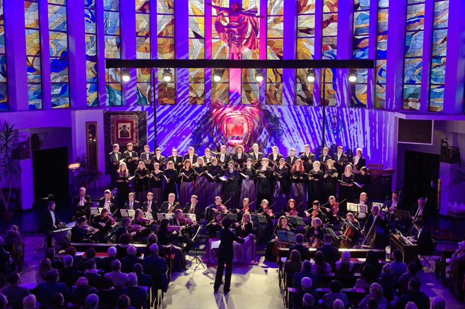 Zdjęcie przedstawia występ zespołu chóralno-instrumentalnego pod batutą Agnieszki Franków-Żelazny podczas koncertu na Festiwalu Gdynia Classica Nova w efektownie oświetlonym wnętrzu z witrażami i liczną publicznością. Chórzyści i muzycy tworzą zwartą, elegancką oprawę muzyczną na tle kolorowej aranżacji świetlnej