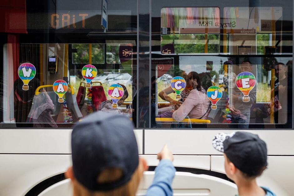 Okna tramwaju oklejone nalepkami tworzącymi napis wakacje . Na pierwszym planie widziane od tyłu postaci dzieci. jedno z nich pokazuje na tramwaj palcem.