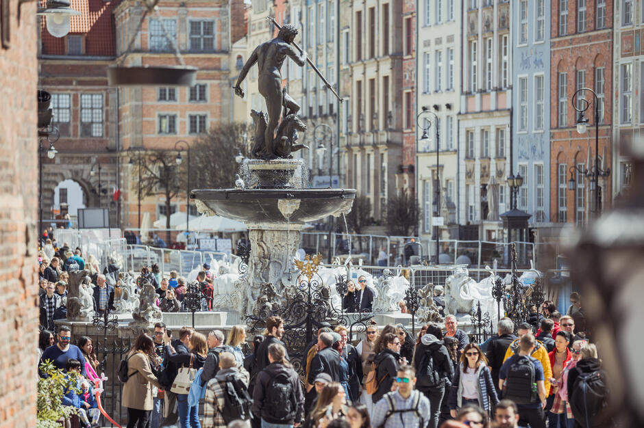 Zdjęcie przedstawia tłum spacerujących turystów wokół Fontanny Neptuna na Długim Targu w Gdańsku, w słoneczny dzień. W tle widać kolorowe, zabytkowe kamienice oraz fragment Zielonej Bramy