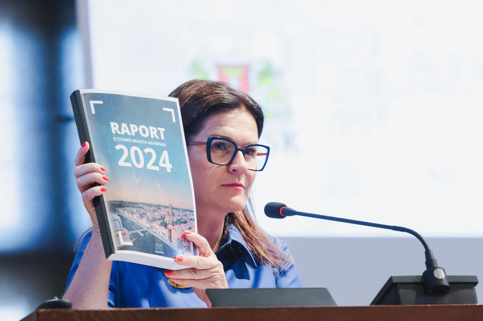 na zdjęciu kobieta w okularach, stoi przy mównicy, trzyma w rękach księgę z napisem raport 2024