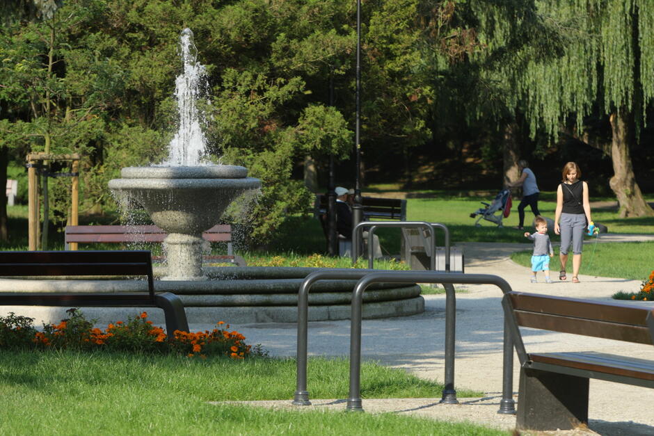 Zdjęcie przedstawia park z fontanną pośrodku, otoczoną kwiatami i ławkami, a w tle widać spacerujących ludzi. Na ścieżce obok fontanny kobieta spaceruje z małym dzieckiem, trzymając je za rękę.