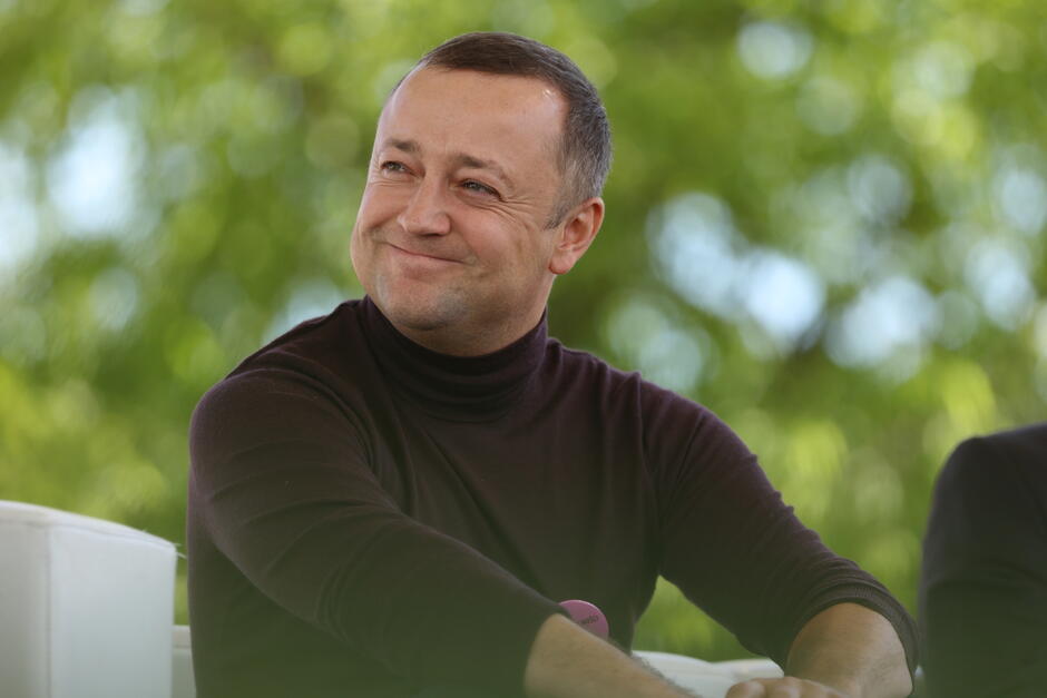 Zdjęcie przedstawia uśmiechniętego mężczyznę w ciemnym golfie, siedzącego na tle zielonych, rozmytych drzew. Mężczyzna patrzy w bok z wyrazem zadowolenia na twarzy, sprawiając wrażenie, jakby uczestniczył w rozmowie lub wydarzeniu na świeżym powietrzu