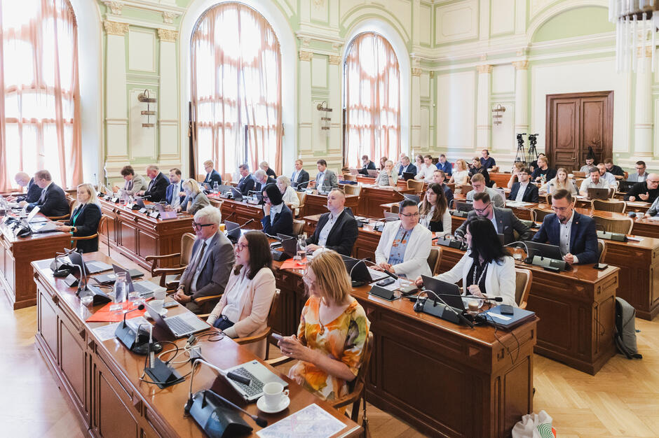 na zdjęciu radni miejscy, siedzą za drewnianymi ławami ustawionymi w kilku rzędach