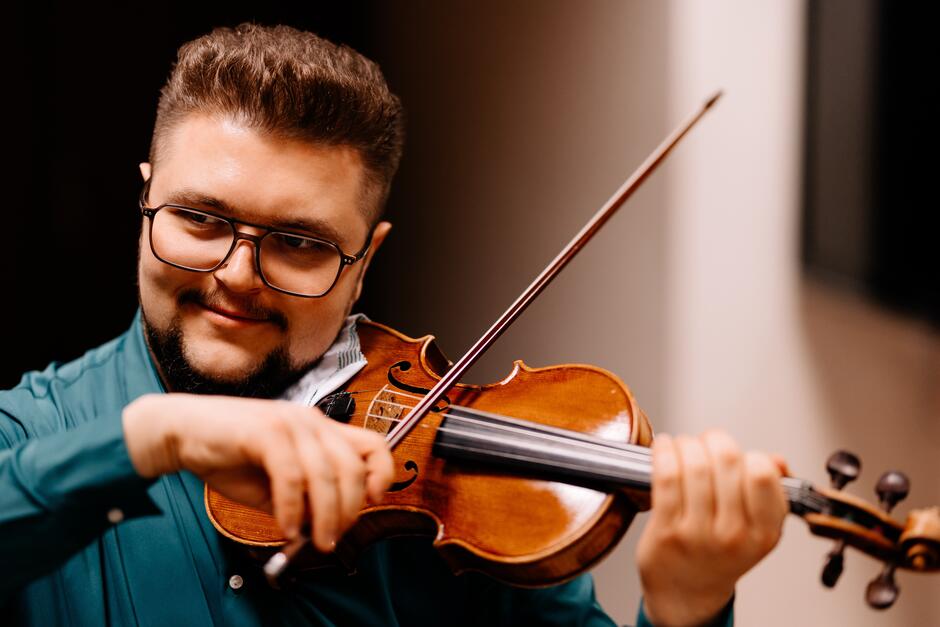 Na zdjęciu mężczyzna w okularach, z krótkimi włosami i brodą, grający na skrzypcach.