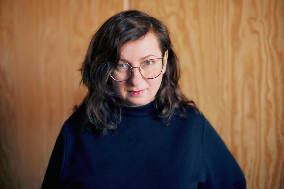 Na zdjęciu kobieta z ciemnymi włosami za ramiona, patrząca na wprost, w tle ściana.