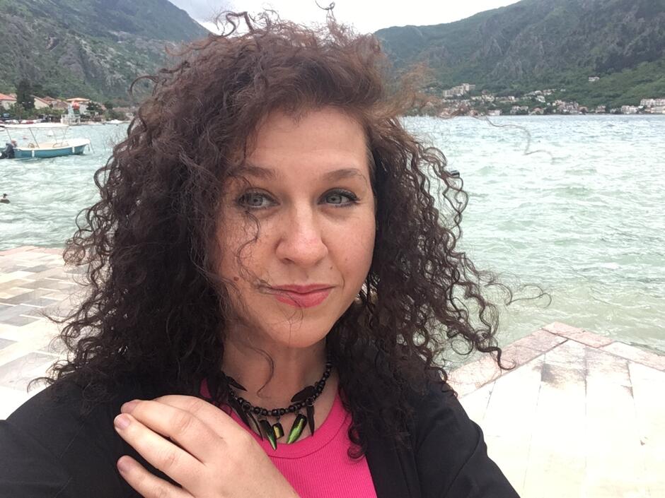 Na zdjęciu kobieta z kręconymi włosami, w tle góry i morze.