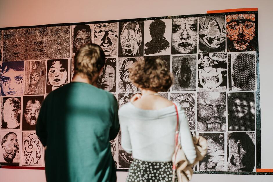 Zdjęcie ukazuje dwie osoby oglądające dużą ścienną kompozycję złożoną z czarno-białych portretów o różnych stylach graficznych. Całość utrzymana jest w kontrastowej estetyce, skupiając uwagę na twarzach i ekspresji