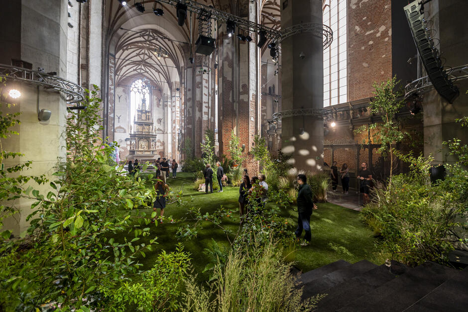 Wnętrze gotyckiego kościoła zostało zamienione w zieloną przestrzeń przypominającą las, z licznymi roślinami i trawą pokrywającą posadzkę. Ludzie spacerują i rozmawiają między drzewkami, a nad całością zawieszono sceniczne oświetlenie