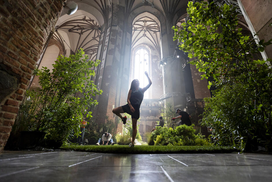 Widok z góry pokazuje całą na zielono zaaranżowaną nawę kościoła, w której ludzie spacerują po trawie wśród roślin i krzewów. Na kolumnach i pod sufitem zamontowano liczne reflektory oraz konstrukcje sceniczne, podkreślające teatralny charakter aranżacji