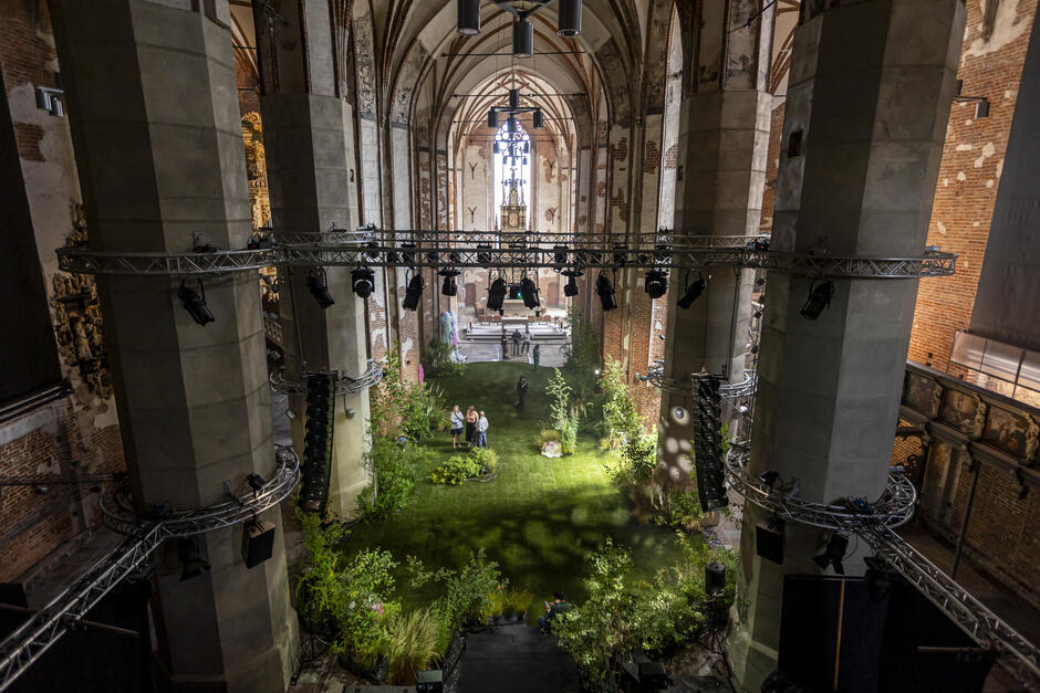 Na tle wysokich, gotyckich okien i łuków sklepienia, kobieta w ciemnej sukience wykonuje taneczny gest w promieniu światła. Wokół niej widać bujną zieleń i osoby pracujące przy aranżacji roślin