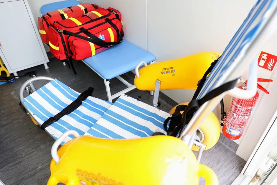 Na zdjęciu znajduje się wnętrze punktu ratowniczego lub medycznego na plaży, wyposażone w specjalistyczny wózek do transportu osób niepełnosprawnych po piasku, oznaczony żółtymi elementami i pasami bezpieczeństwa. W tle widoczna jest czerwona torba ratownicza, niebieska leżanka oraz gaśnica zamocowana na ścianie. 