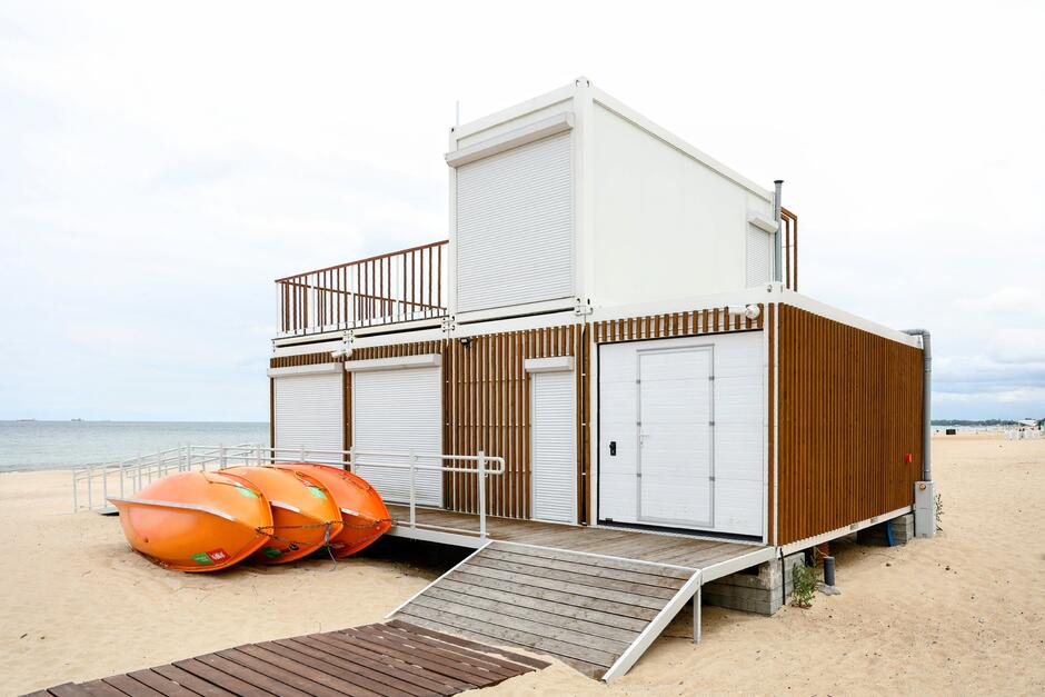 Na zdjęciu widać nowoczesny budynek plażowy wykonany z kontenerów, wykończony drewnianymi panelami i wyposażony w rolety zamykające okna oraz drzwi. Obok niego znajdują się trzy pomarańczowe kajaki ułożone na piasku, a całość położona jest tuż przy brzegu morza. 