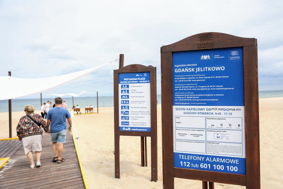 Na zdjęciu widoczna jest wejściowa strefa plaży Gdańsk Jelitkowo, z drewnianymi chodnikami prowadzącymi na piasek oraz tablicami informacyjnymi dotyczącymi kąpieliska i udogodnień dla plażowiczów. W tle widać morze, kilka osób spacerujących oraz rozciągnięte nad chodnikiem białe żagle dające cień. 