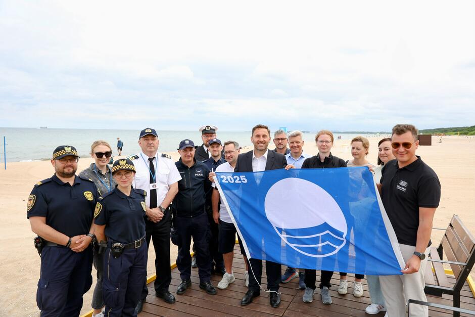 Na zdjęciu widoczna jest grupa osób, w tym przedstawiciele służb mundurowych, urzędnicy i pracownicy plaży, pozująca na tle morza na plaży w Gdańsku Jelitkowie. Trzymają oni niebieską flagę programu „Blue Flag 2025”, symbolizującą wysoką jakość kąpieliska i czystość środowiska. 
