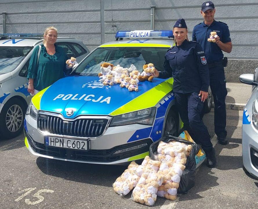 Na zdjęciu widzimy dwóch policjantów oraz kobietę stojących obok radiowozu oznaczonego napisem „Policja”, na którego masce i przed nim ułożone są liczne pluszowe misie. Pluszaki prawdopodobnie zostaną rozdane dzieciom w ramach akcji wsparcia najmłodszych w trudnych sytuacjach.
