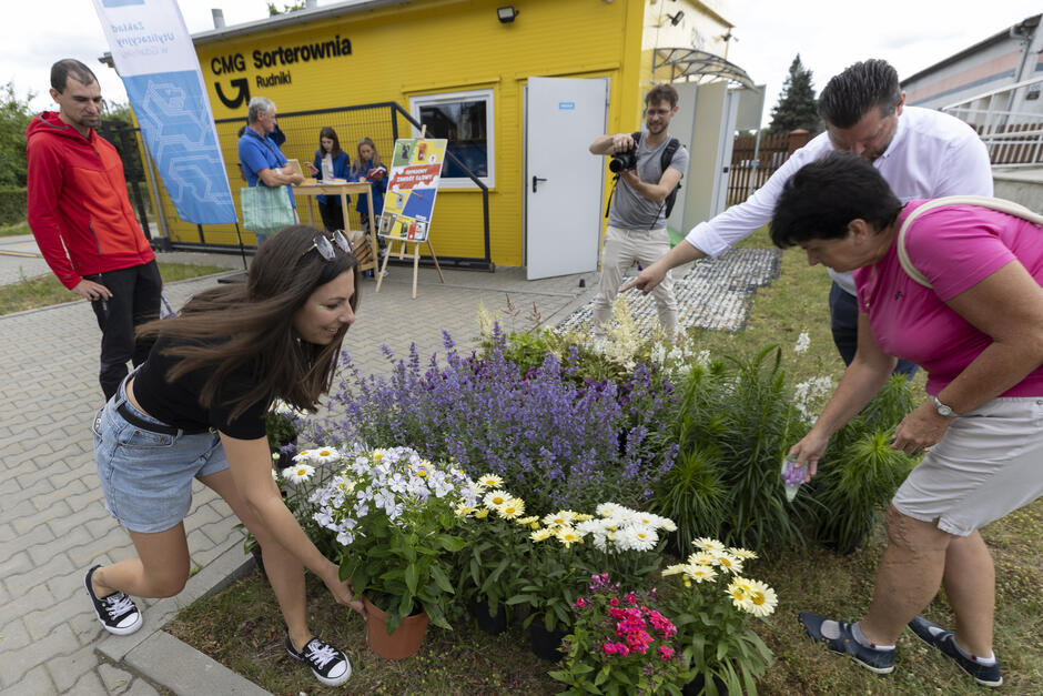na zdjęciu młoda kobieta pochyla się nad kwiatami w doniczkach, po drugiej stronie pochyla się druga kobieta, w tle dwóch mężczyzn przygląda się im