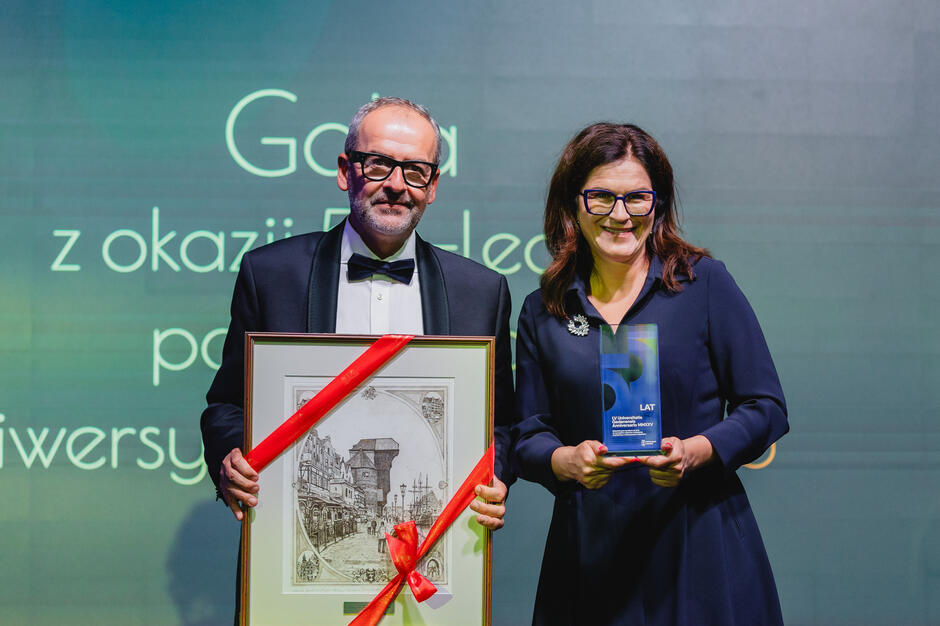 na zdjęciu z lewej starszy mężczyzna w smokingu, pozuje do zdjęcia z dużym oszklonym obrazem, obok kobieta w okularach w średnim wieku, uśmiecha się